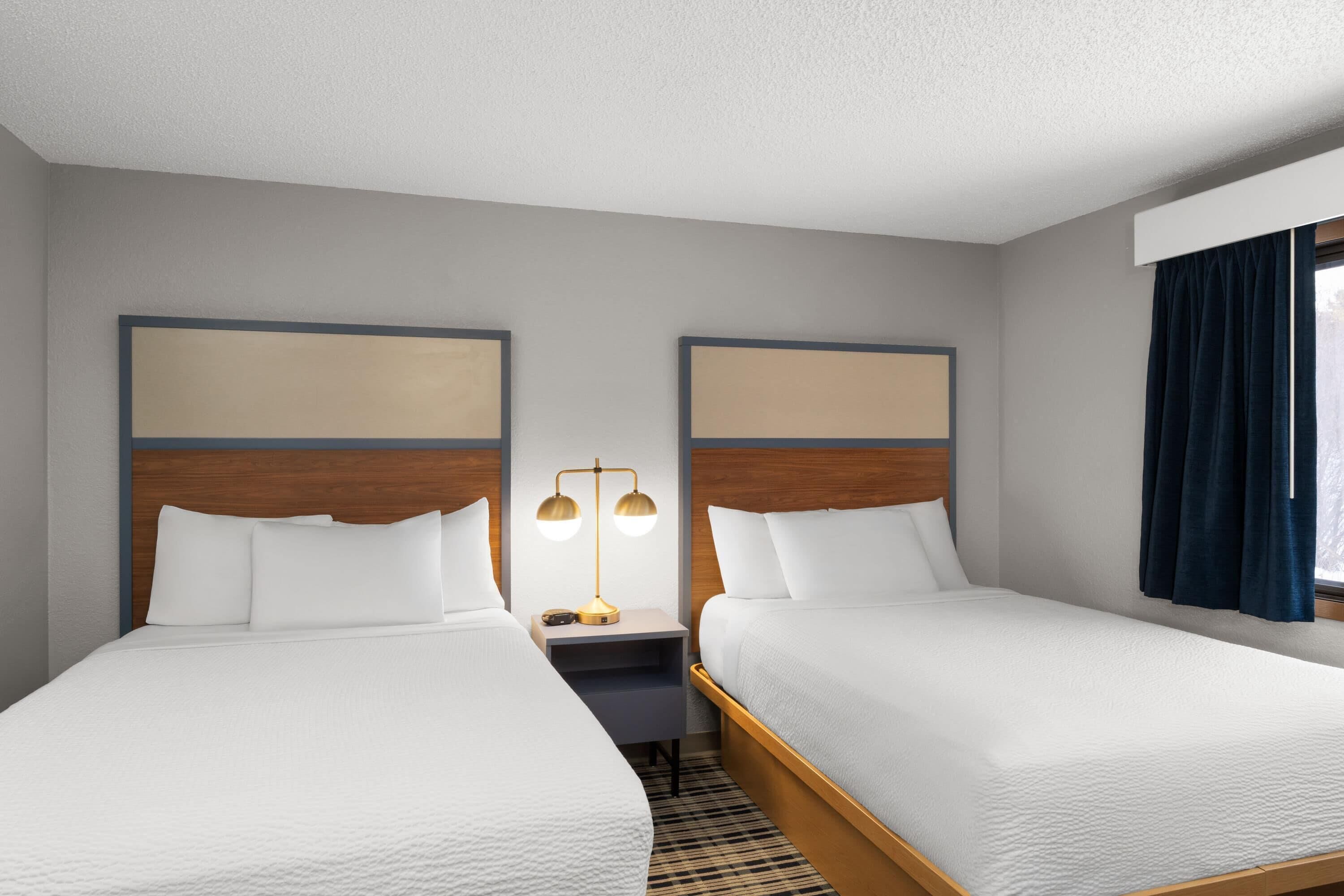 A hotel room with two single beds, a nightstand with a lamp, and a window with curtains.