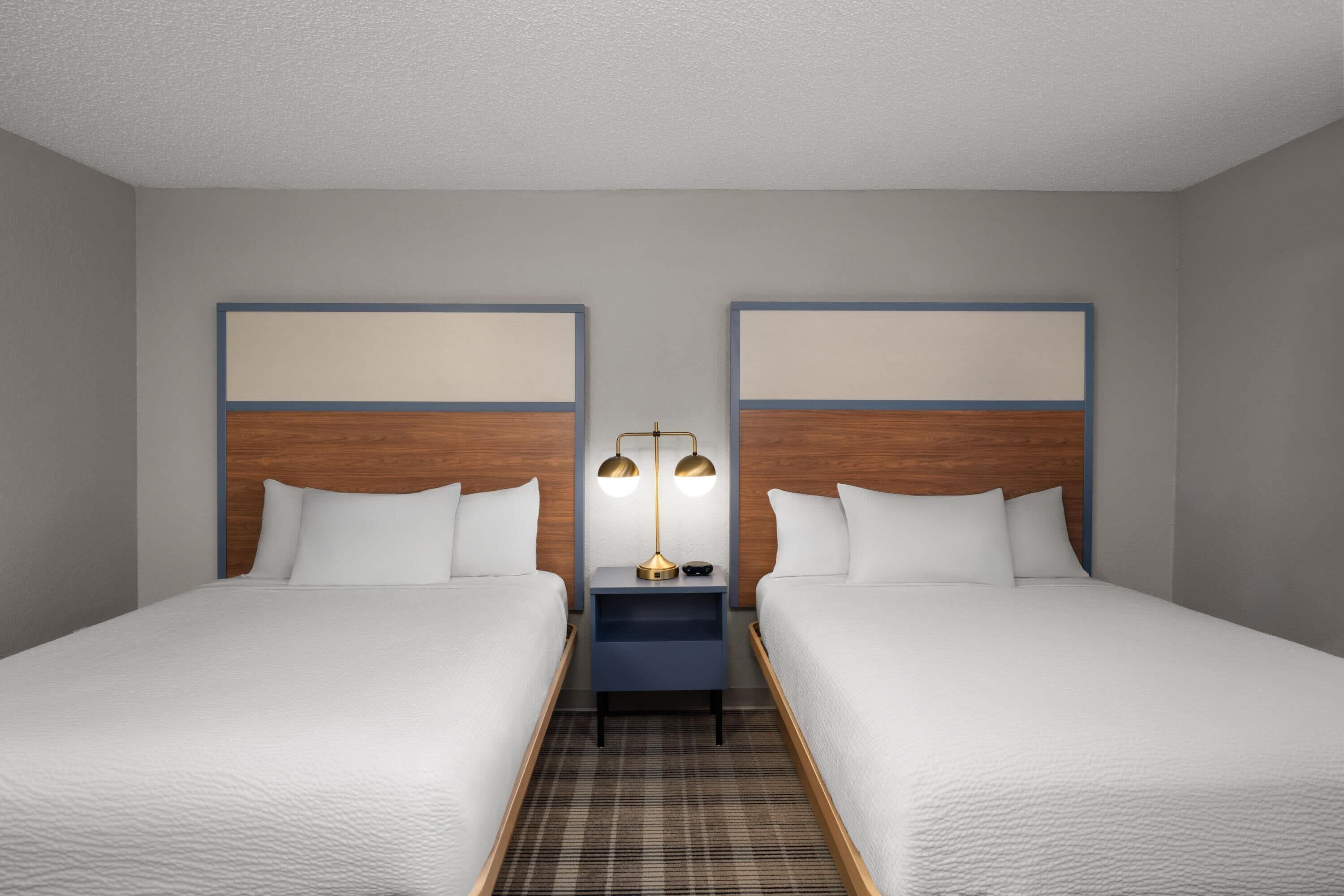 Una habitación de hotel con dos camas, una mesita de noche con lámpara y una alfombra estampada de pata de gallo.