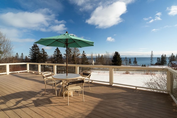 A wooden deck with a table and chairs, a green umbrella, and a view of a snowy landscape and trees.