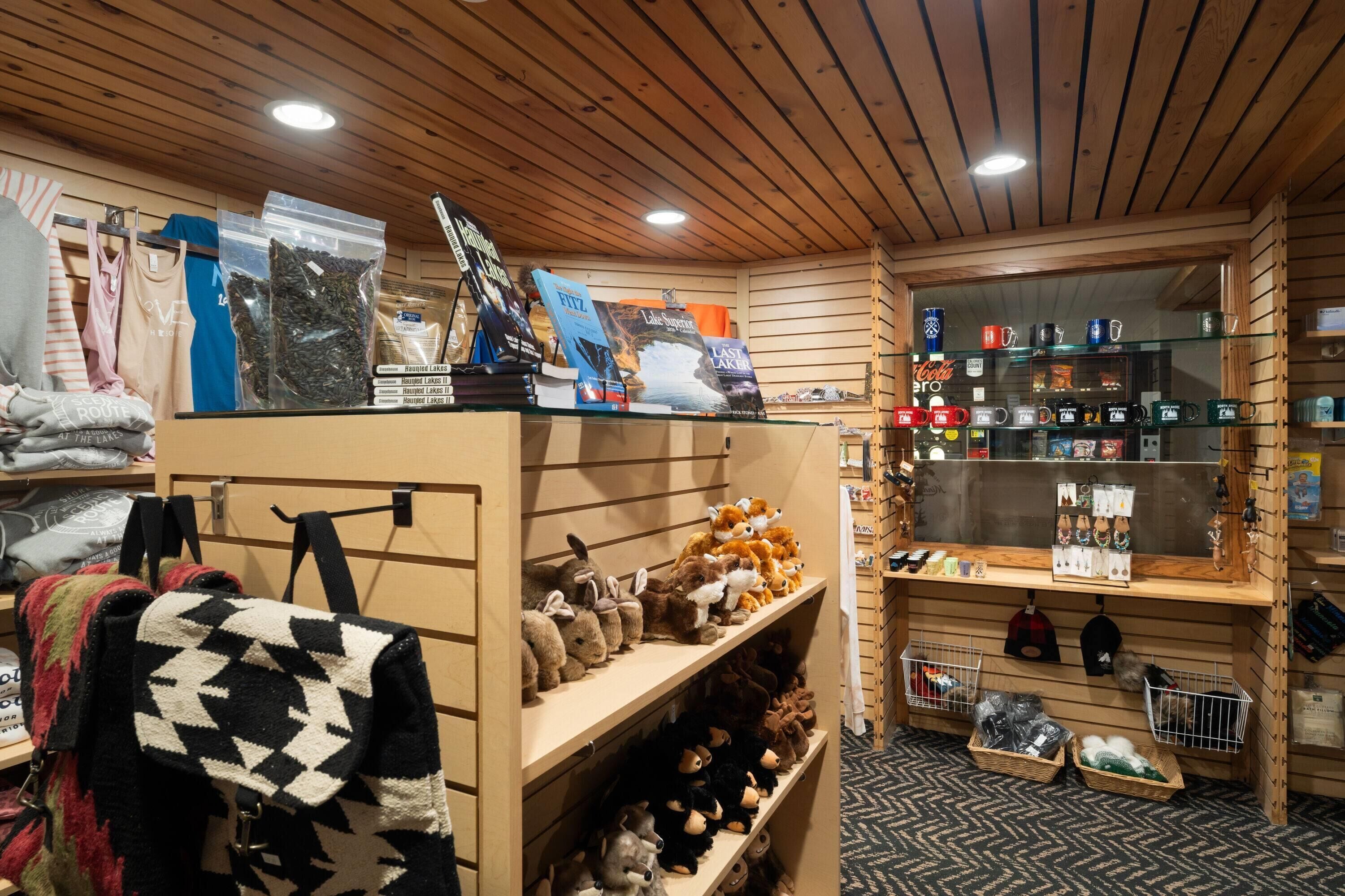 A cozy room with wooden walls and ceiling, shelves with plush toys and bags, and a display of mugs and other items.