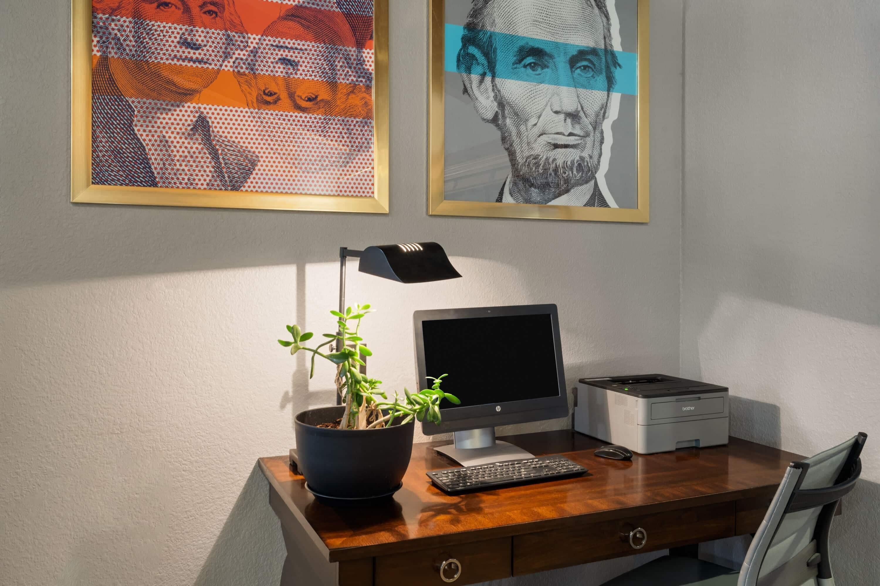 Un bureau en bois avec un ordinateur de bureau, une imprimante, une plante en pot et une œuvre d’art encadrée accrochée au mur.