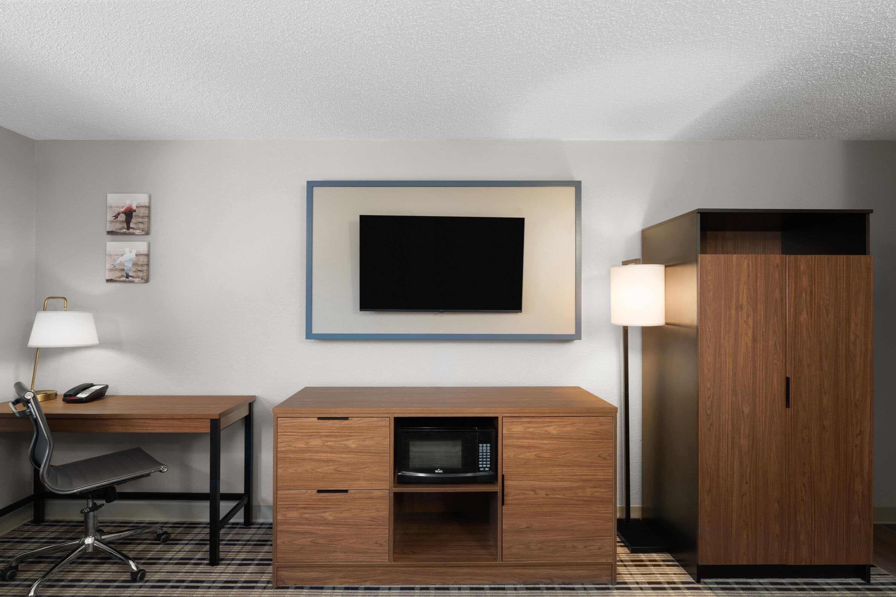 A hotel room with a wooden desk, a chair, a microwave, a TV, a lamp, and a wardrobe.