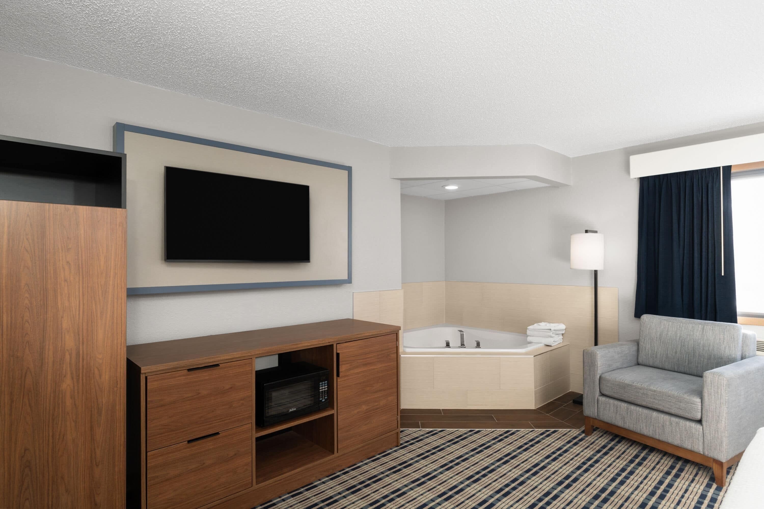 Una habitación de hotel moderna con una televisión de pantalla plana montada en la pared, un mueble de madera con cajones, un sillón gris, una bañera grande y una lámpara de pie.