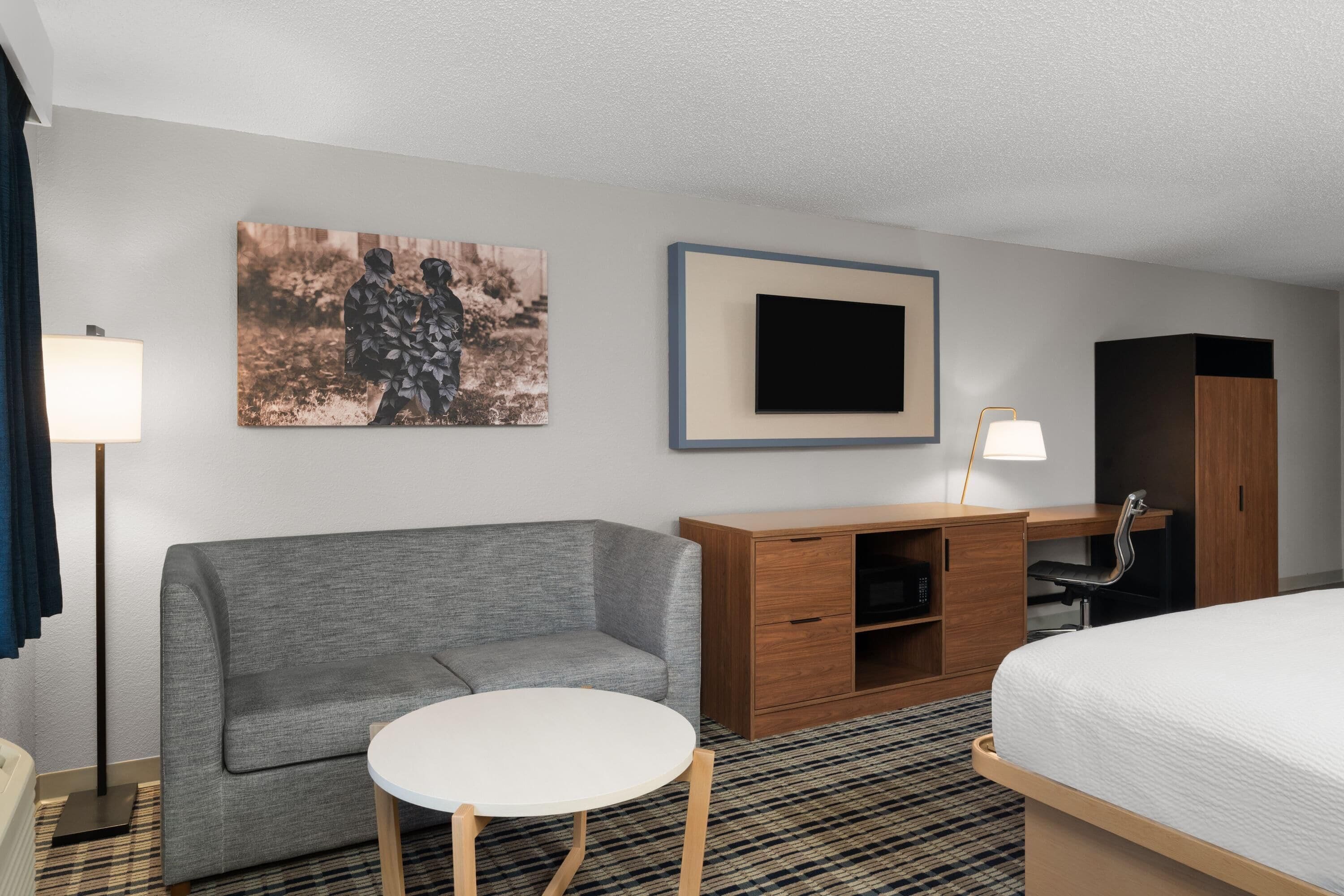 Ein Hotelzimmer mit grauem Sofa, einem weißen runden Tisch, einem Holzbüro mit Stuhl, einem Bett, einem an der Wand montierten Fernseher und einem Gemälde über dem Schreibtisch.