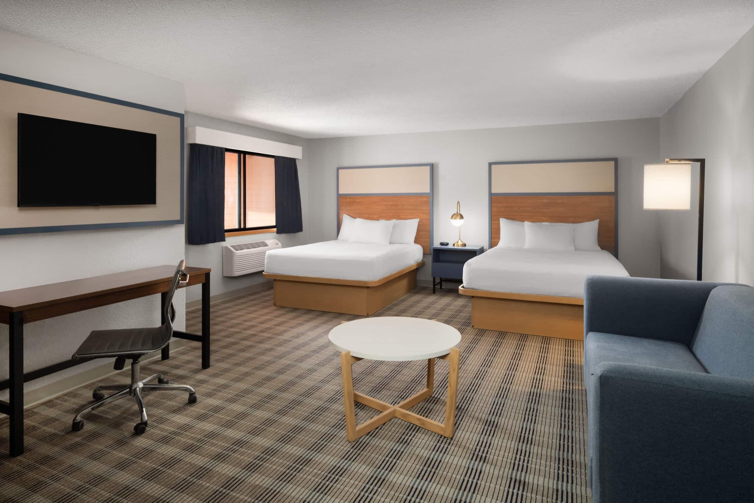 A hotel room with two beds, a desk, a chair, a small table, a TV, and a lamp.