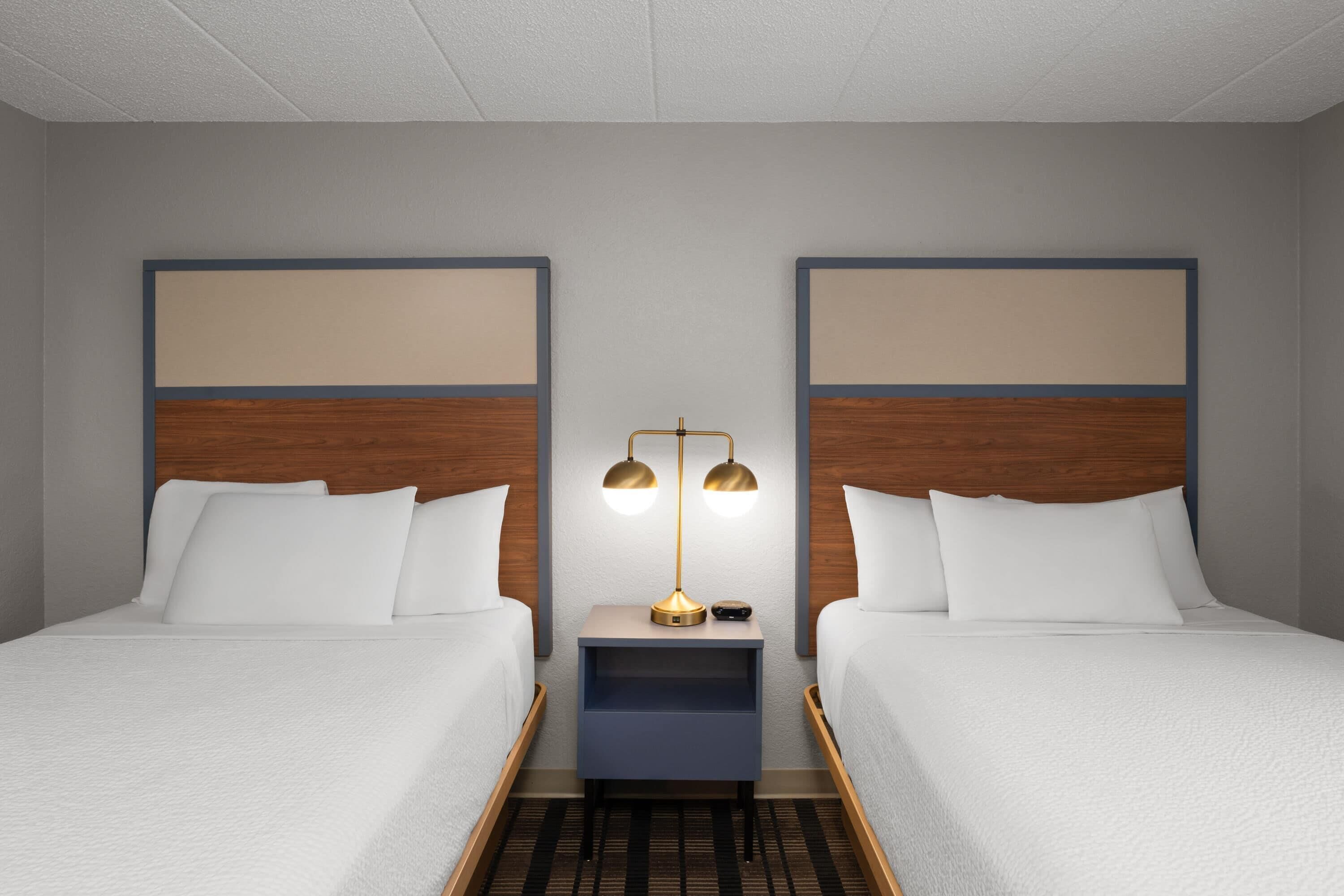 A hotel room with two beds, a nightstand with a lamp, and a headboard with a wooden panel.