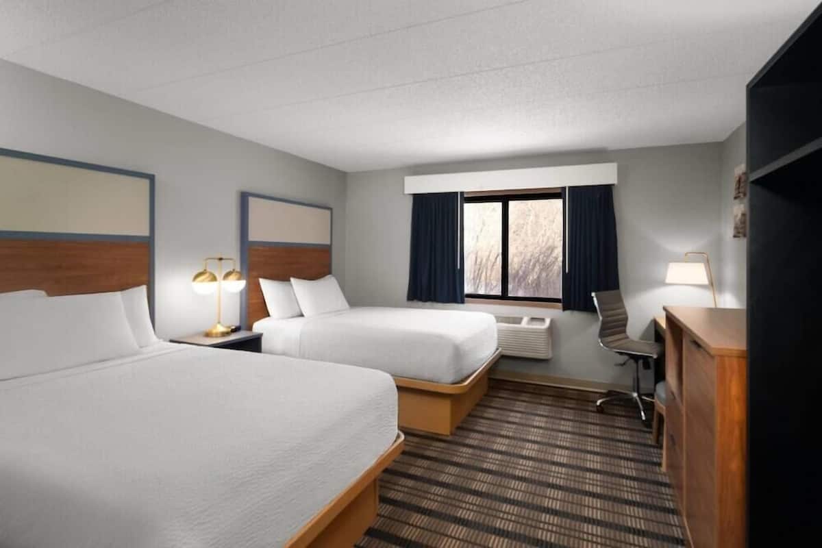 A hotel room with two beds, a desk, a chair, a lamp, and a window with curtains.