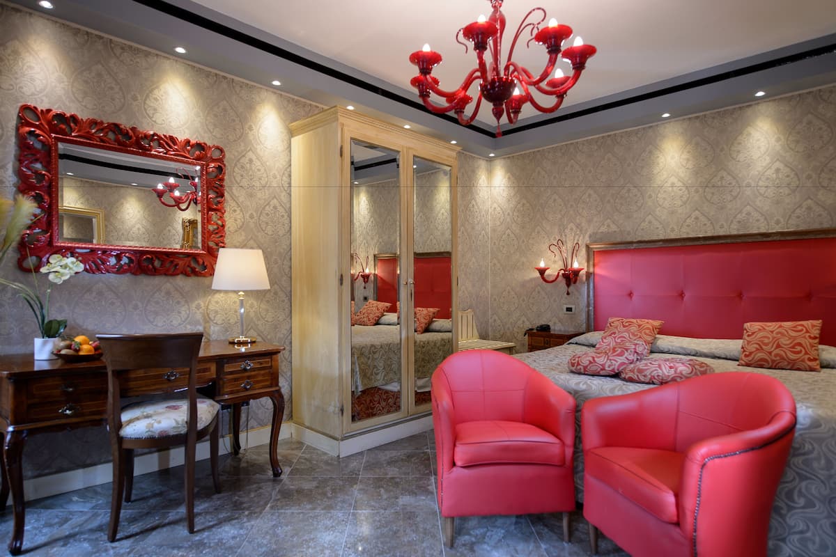 Un dormitorio con una cama grande, cabecera roja, espejo con marco rojo, escritorio con silla y sillón rojo.