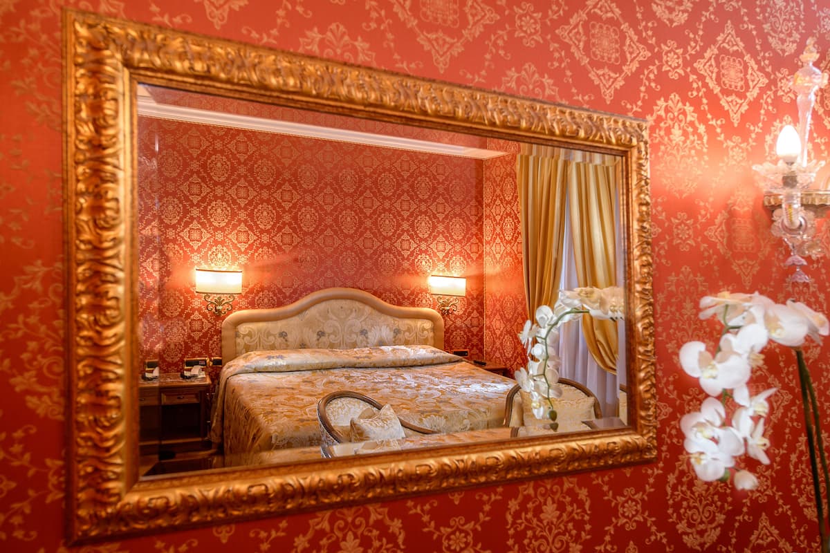 Una habitación con un espejo grande y ornamentado, una cama con cabecera, dos lámparas de noche y un arreglo floral.