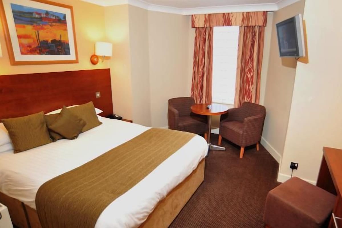 A hotel room with a large bed, two armchairs, a small table, a TV, and a painting on the wall.