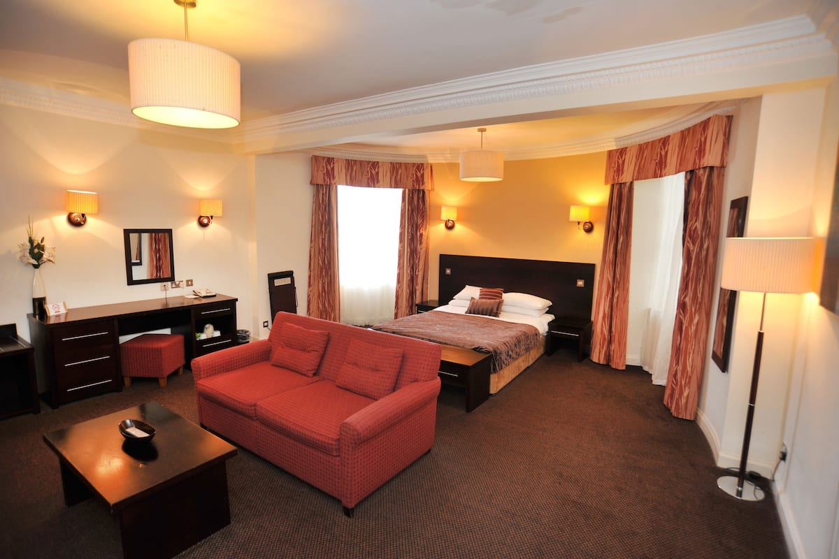 A hotel room with a bed, a red sofa, a desk, and a lamp.