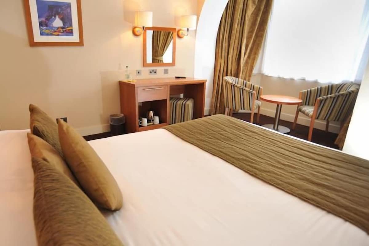 Une chambre d’hôtel avec un grand lit, une table de chevet en bois, un miroir, une petite table avec des chaises et une fenêtre avec des rideaux.