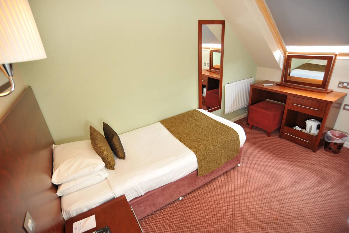 A small, neatly arranged bedroom with a bed, a desk, a chair, and a mirror.