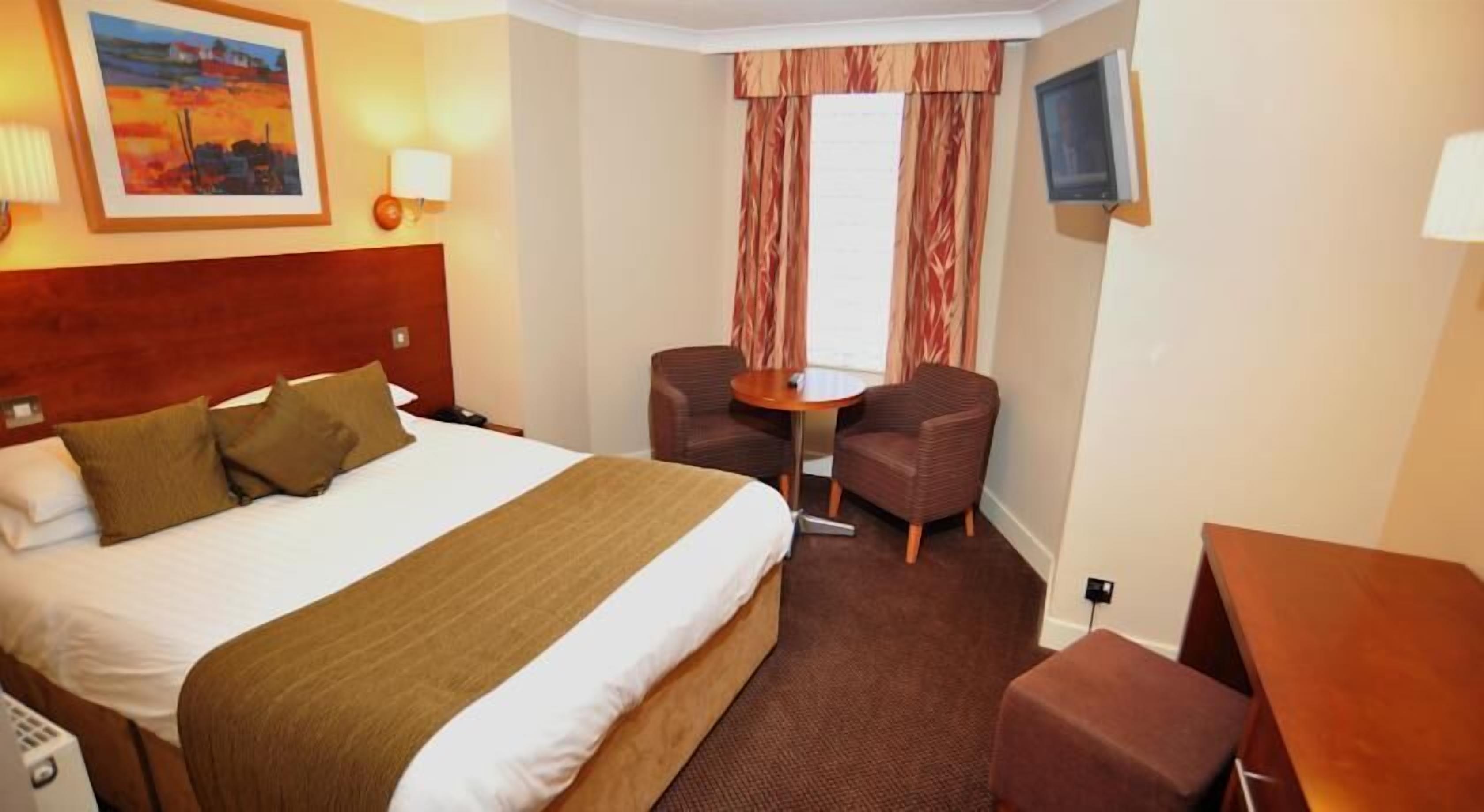 A hotel room with a large bed, two armchairs, a small table, a TV, and a painting on the wall.