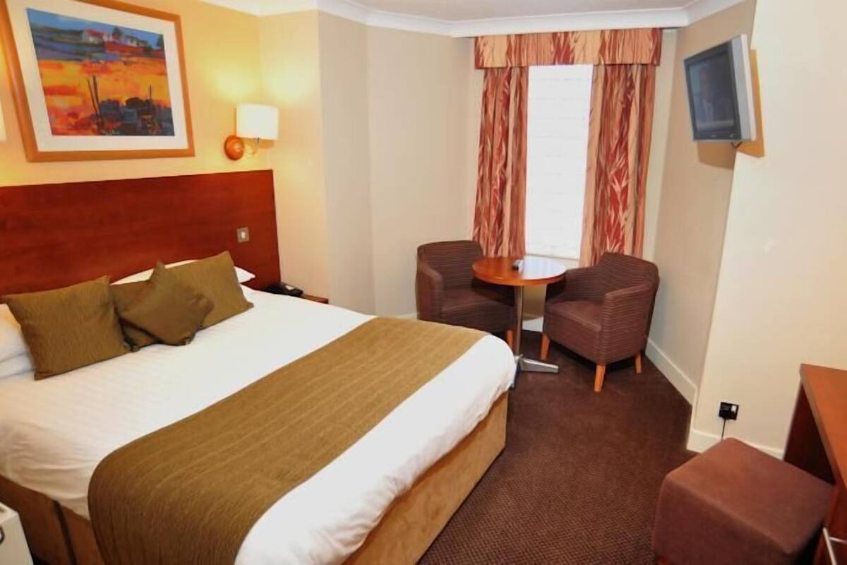A hotel room with a large bed, two armchairs, a small table, a TV, and a painting on the wall.