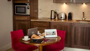 A kitchen with wooden cabinets, a microwave, a kettle, and a sink. There is a table with a laptop, a cup, and pastries.