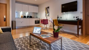 A modern living room with a grey sofa, a wooden coffee table with magazines and a tablet, a red chair, and a yellow chair. A kitchen with white cabinets and a TV is in the background. - Premier Suites Dublin Leeson Street (Dublin)