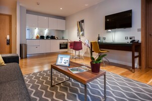 A modern living room with a grey sofa, a wooden coffee table with magazines and a tablet, a red chair, and a yellow chair. A kitchen with white cabinets and a TV is in the background.