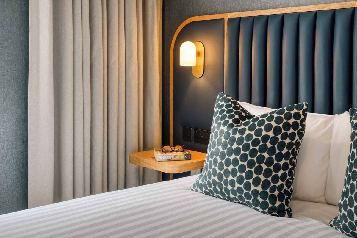 A hotel room with a bed, a nightstand with glasses and a book, a wall-mounted light, and a window with curtains.