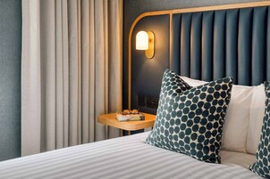 A hotel room with a bed, a nightstand with glasses and a book, a wall-mounted light, and a window with curtains.