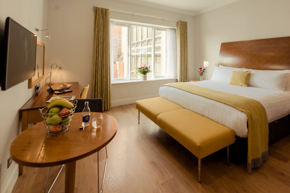 A hotel room with a bed, a desk, a chair, a table with fruits and a bottle, and a window with curtains.