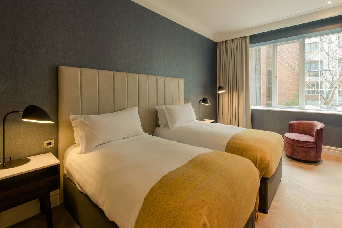 A hotel room with two beds, a nightstand, a chair, and a window with a view of buildings.