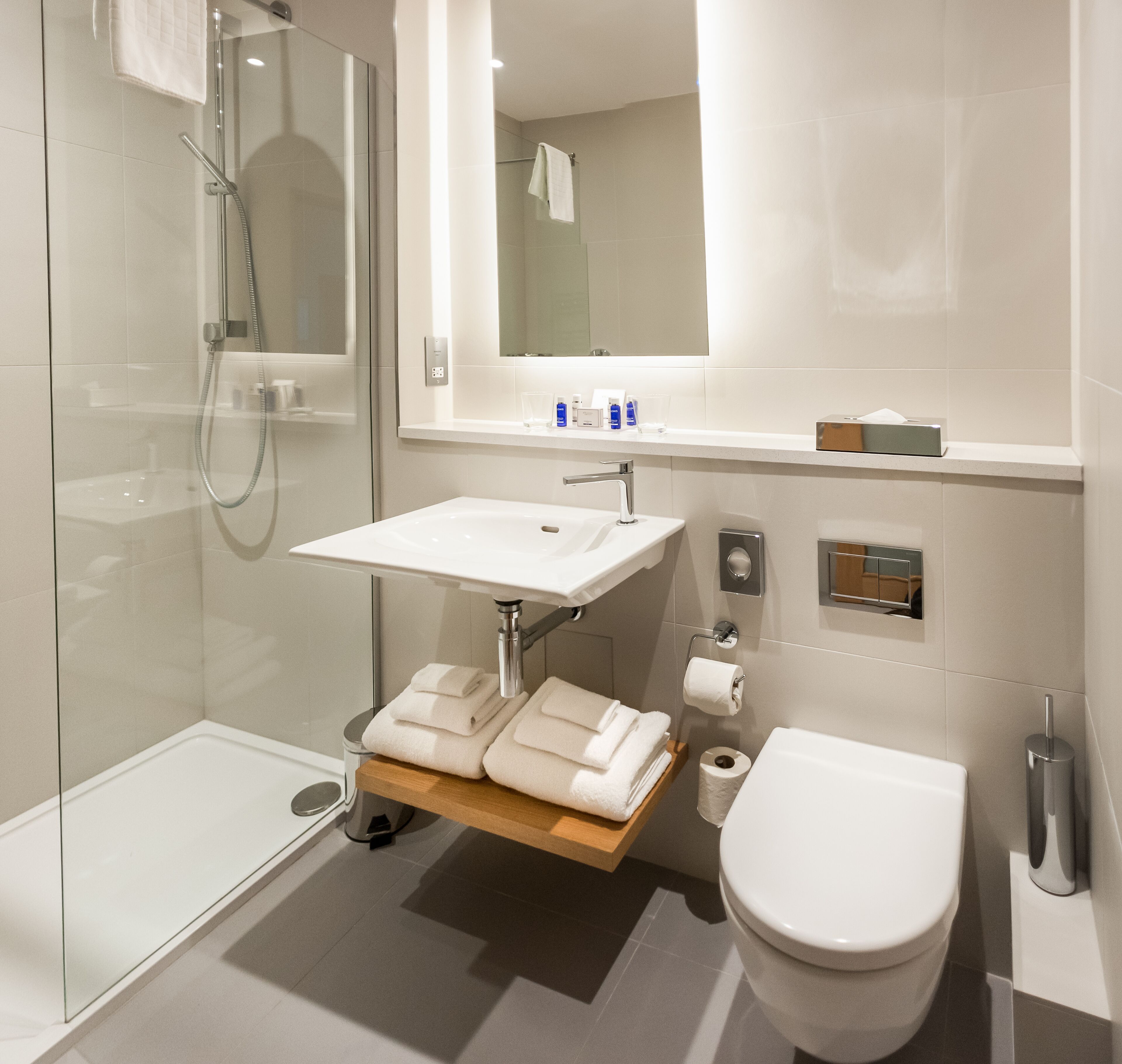Un baño moderno con ducha envuelta en vidrio, un lavamanos blanco con espejo arriba y un inodoro con dispensador de toallas de papel.