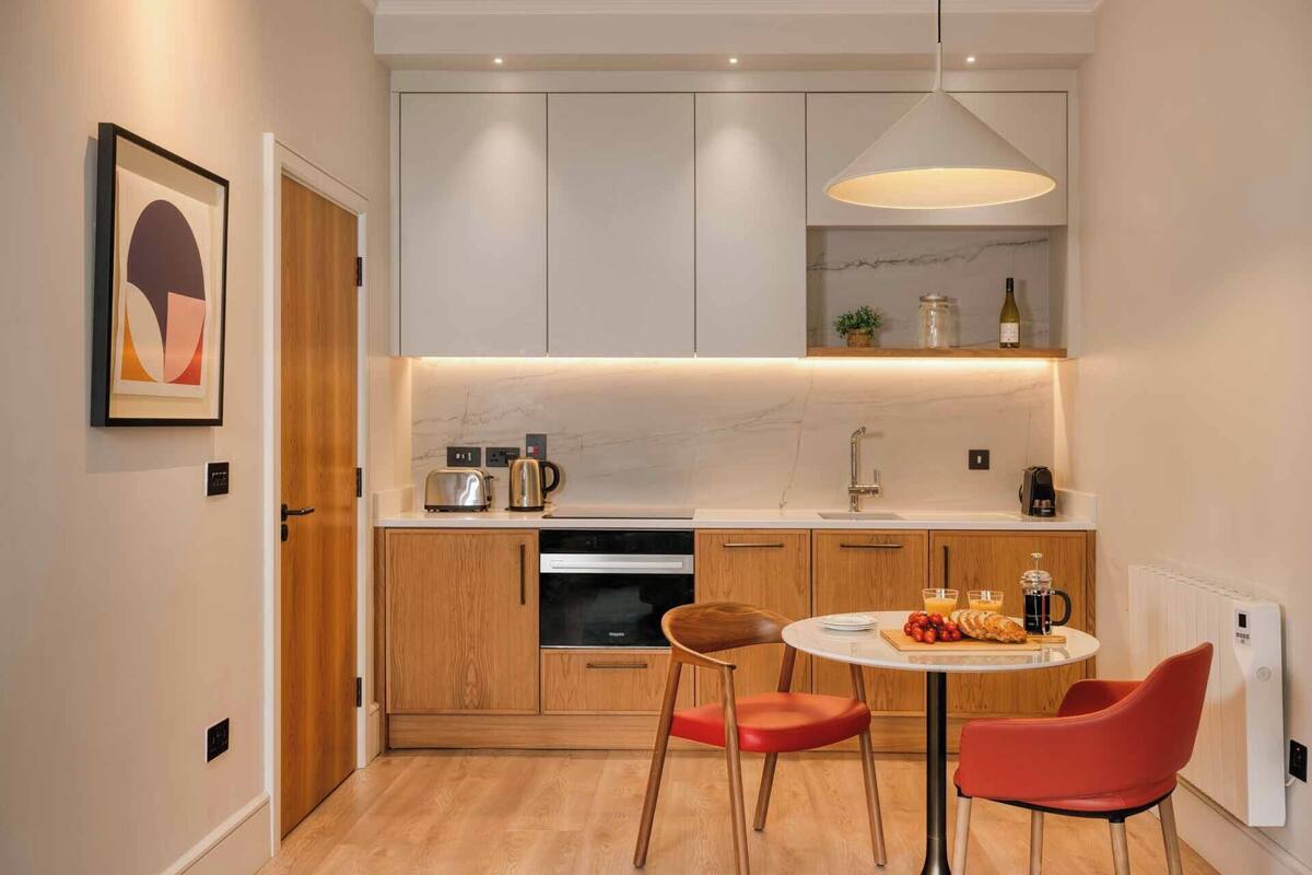 A modern kitchen with wooden cabinets, a built-in oven, a round dining table with two chairs, and a pendant light above the counter.