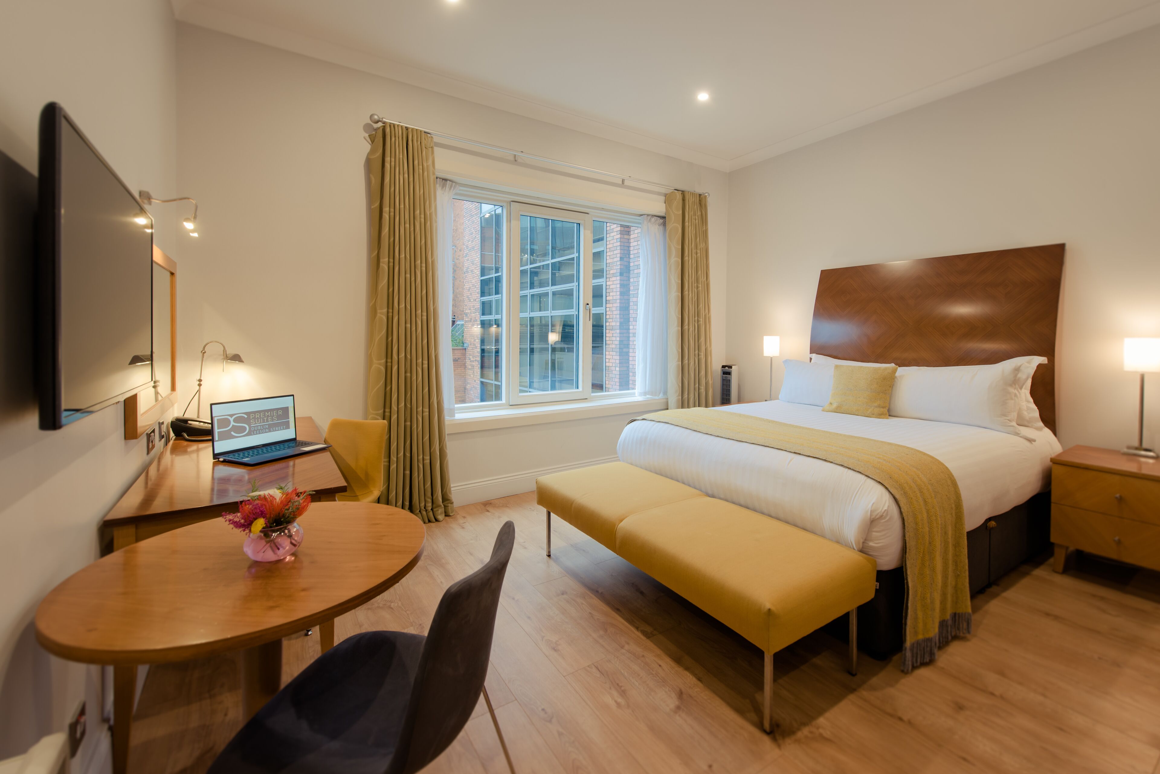 Habitación de hotel con cama, mesita de noche, lámpara, ventana con cortinas, escritorio con una computadora portátil, silla y una pequeña mesa con un jarrón de flores.