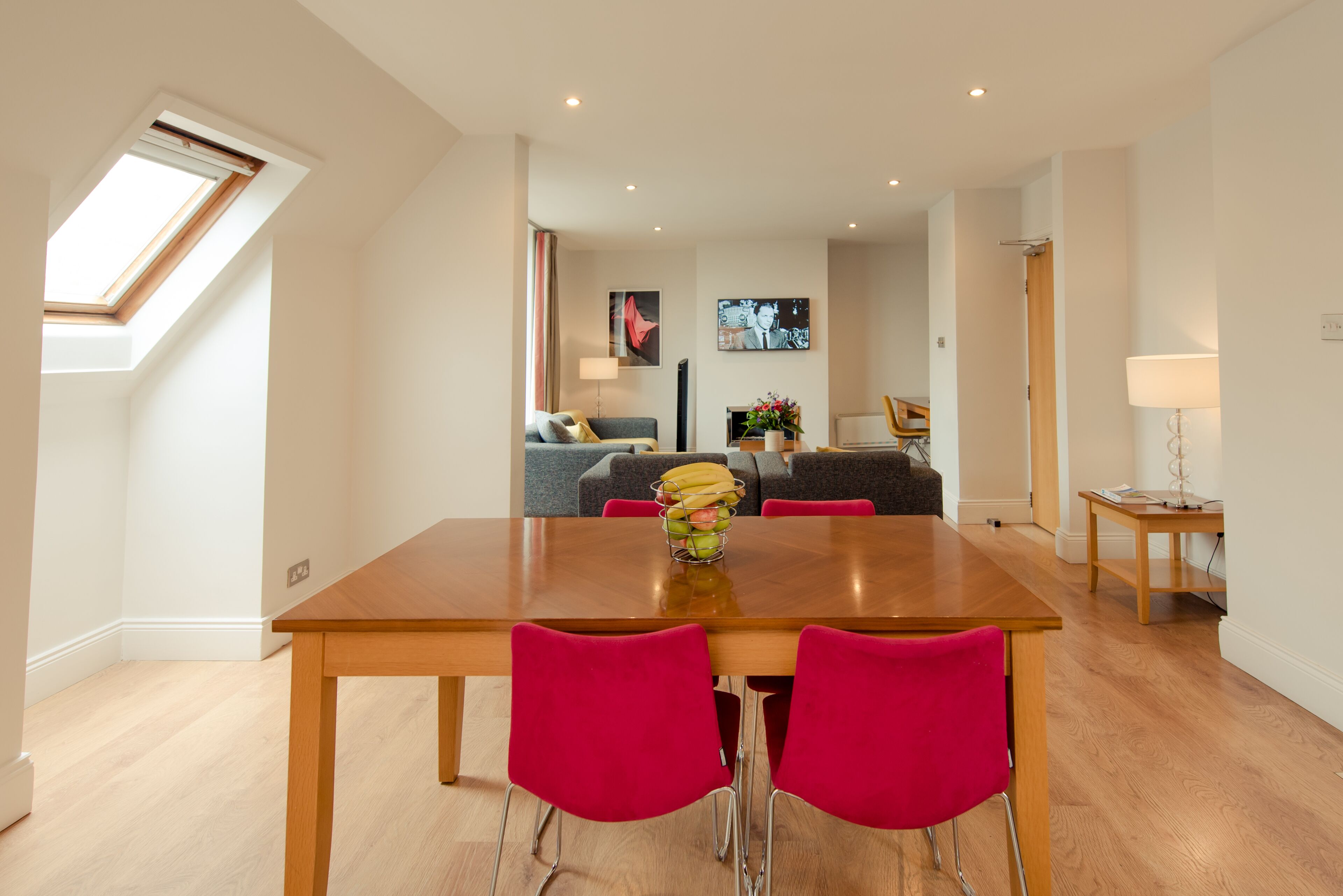 Un comedor moderno con una mesa de madera y sillas rojas, un sofá gris y un televisor al fondo.