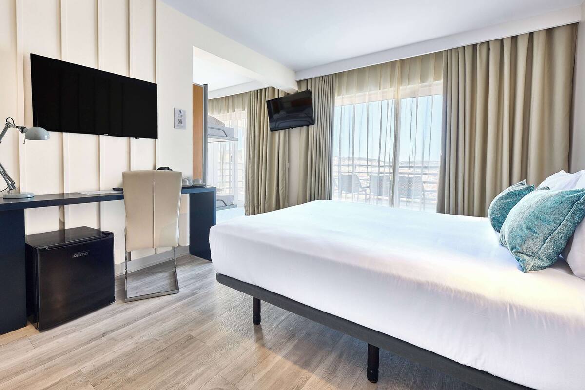 Habitación de hotel con cama, escritorio, silla, televisor montado en la pared y vistas a la ciudad a través de amplias ventanas.