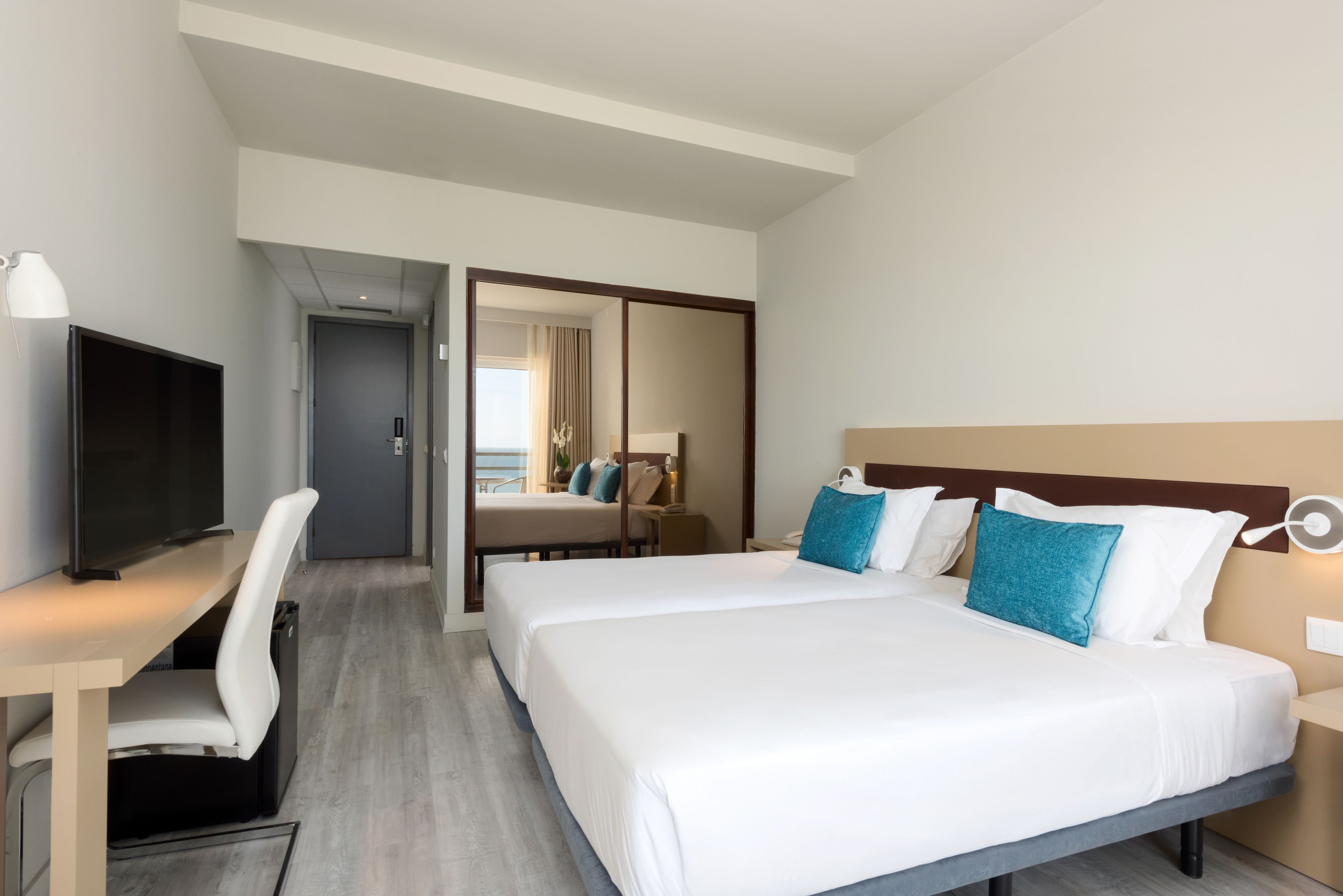 Habitación de hotel con una cama grande, un escritorio con silla, televisión y una puerta que da a otra habitación.