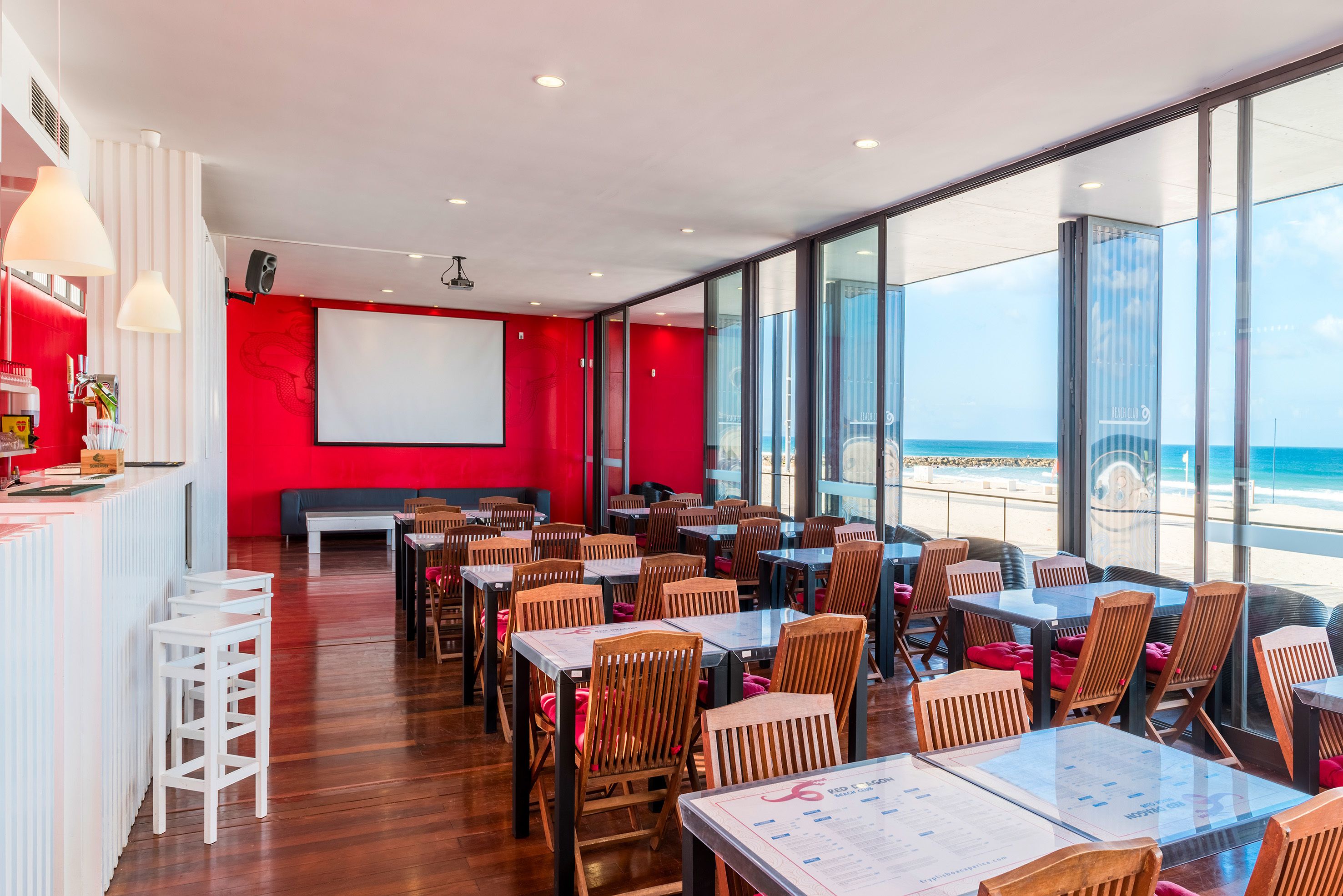 A restaurant with a red interior, wooden tables and chairs, a white counter, and large windows overlooking the beach.