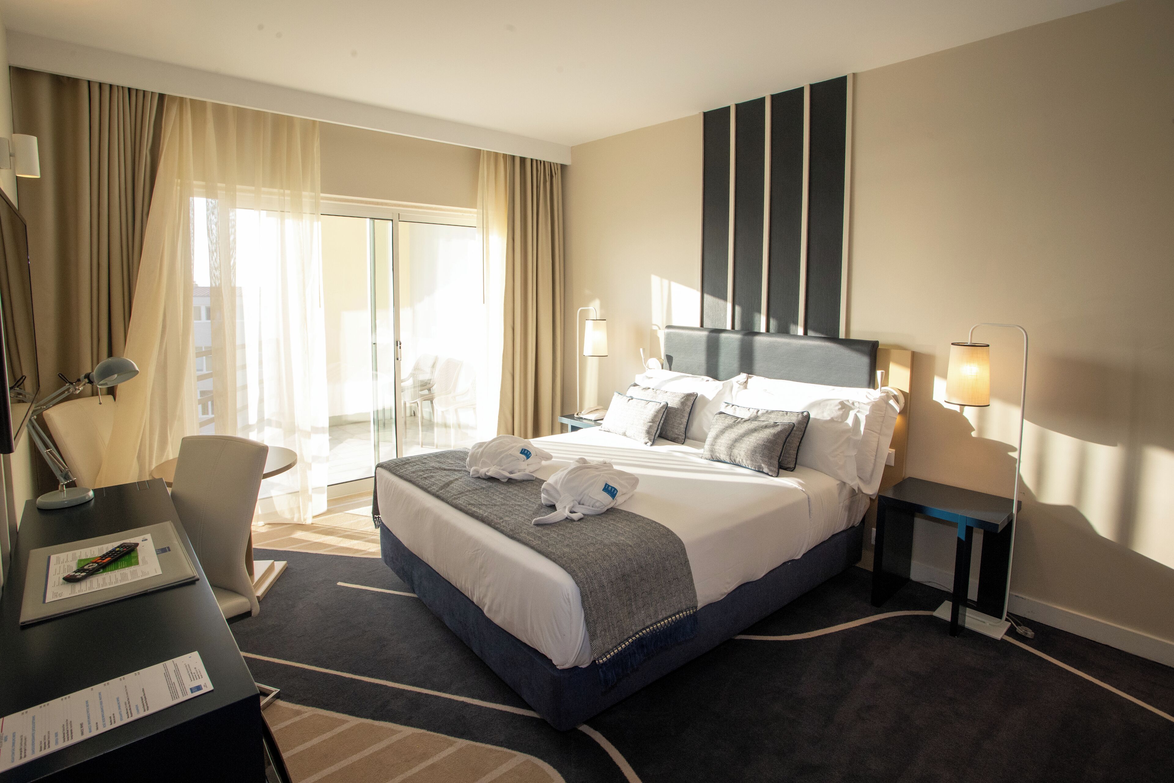 A hotel room with a large bed, a desk with a laptop, a chair, and a view of the outdoors through a sliding door.