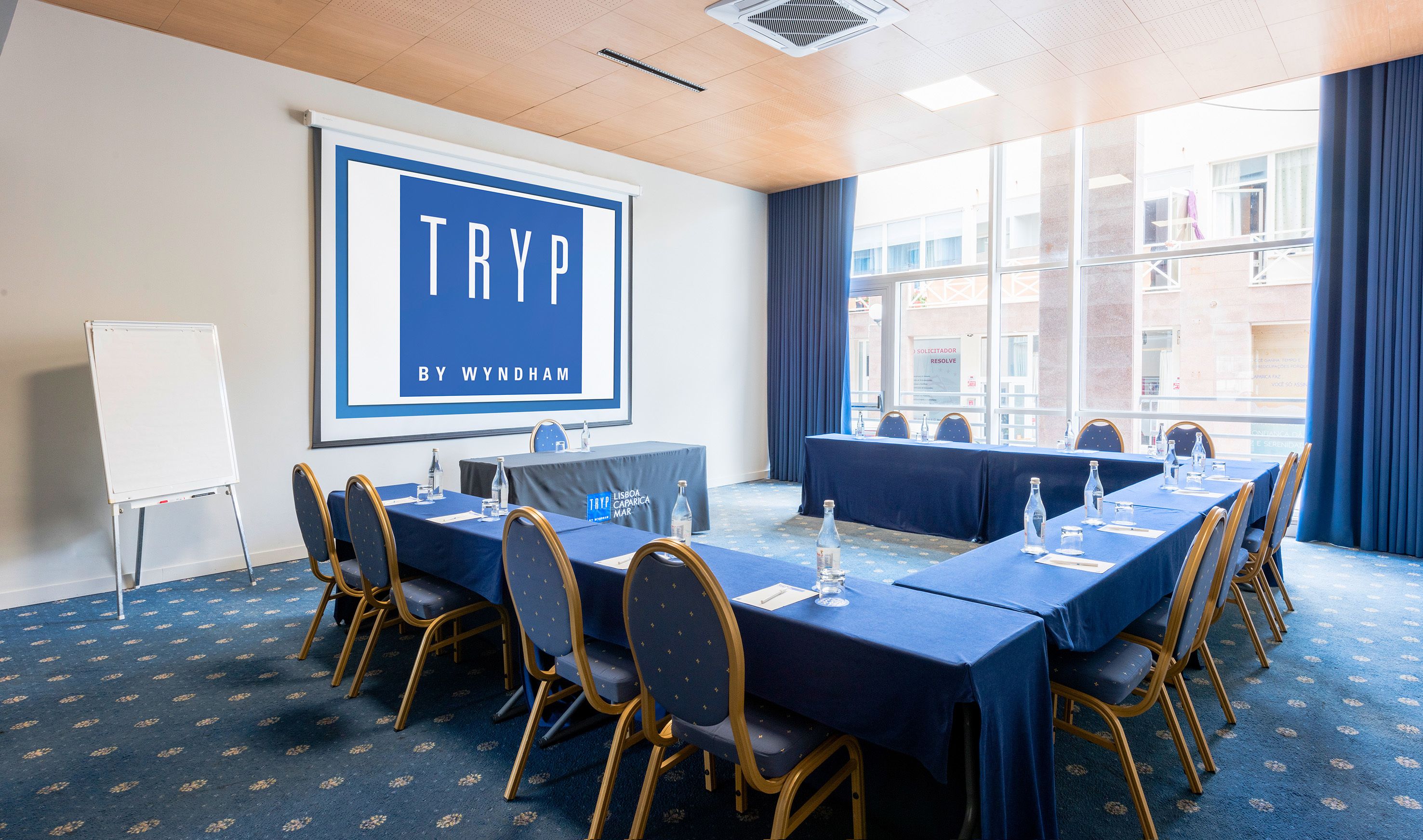A conference room with a large screen displaying "TRYP BY WYNDHAM", a long table with chairs, and a whiteboard.