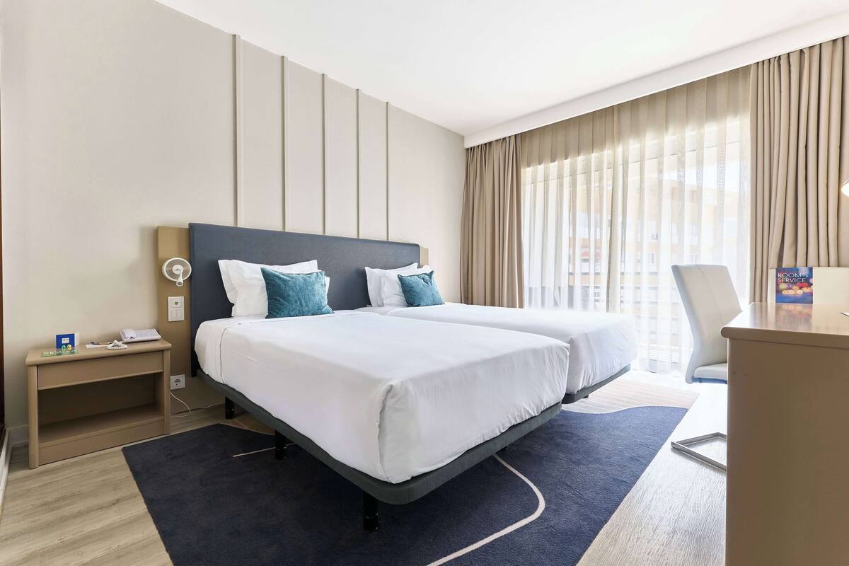 Habitación de hotel con dos camas, un escritorio, una silla y vista de la ciudad a través de amplias ventanas.