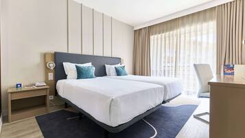 Habitación de hotel con dos camas, un escritorio, una silla y vista de la ciudad a través de amplias ventanas.