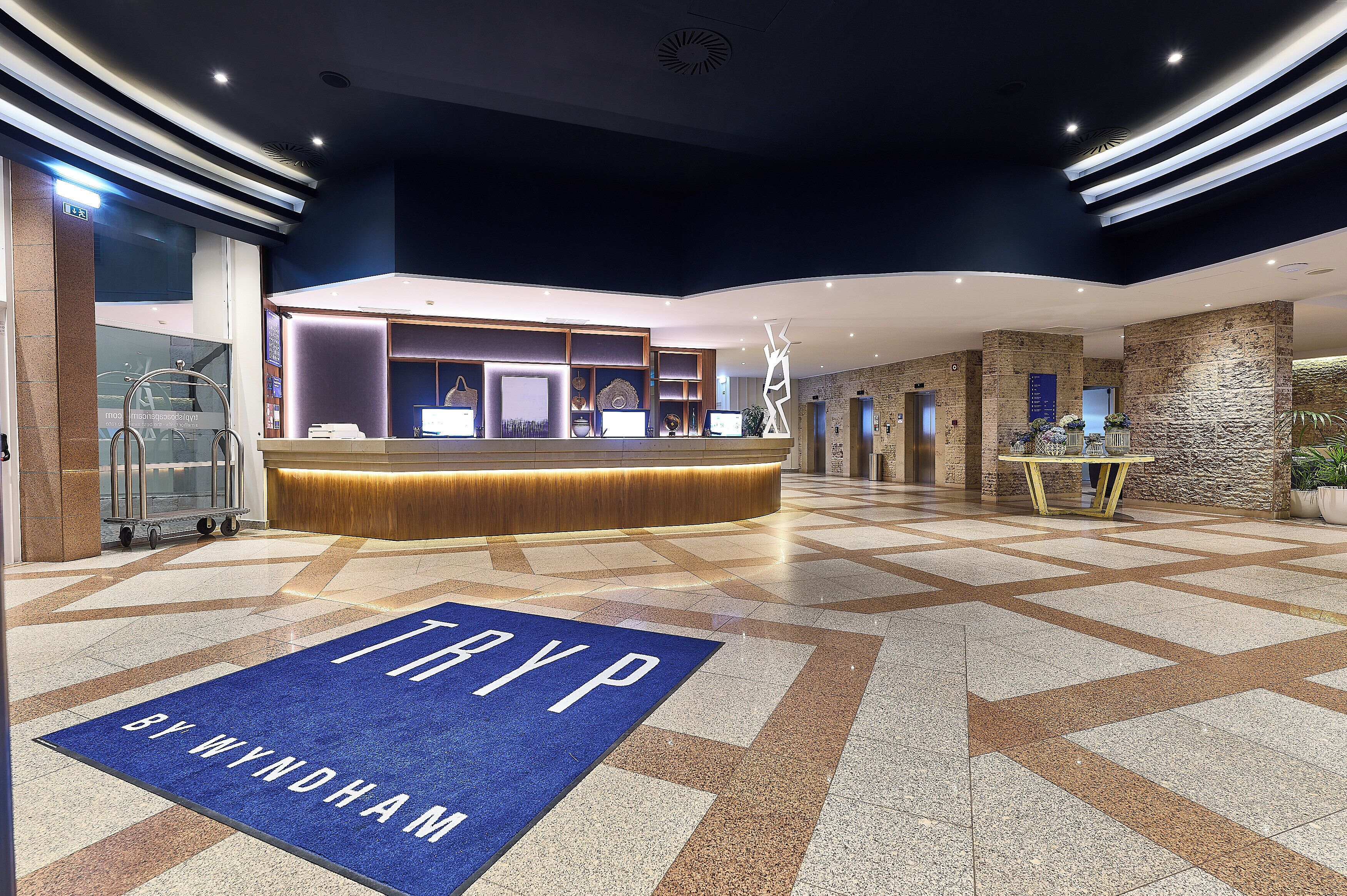 Een moderne hotellobby met een receptiebalie, een blauw tapijt met de tekst 'TRYP BY WYNDHAM' en een geometrisch betegelde vloer.