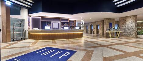 Een moderne hotellobby met een receptiebalie, een blauw tapijt met de tekst 'TRYP BY WYNDHAM' en een geometrisch betegelde vloer.
