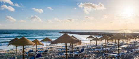 A beach with rows of sun loungers and thatched umbrellas, a calm sea, and a clear sky.