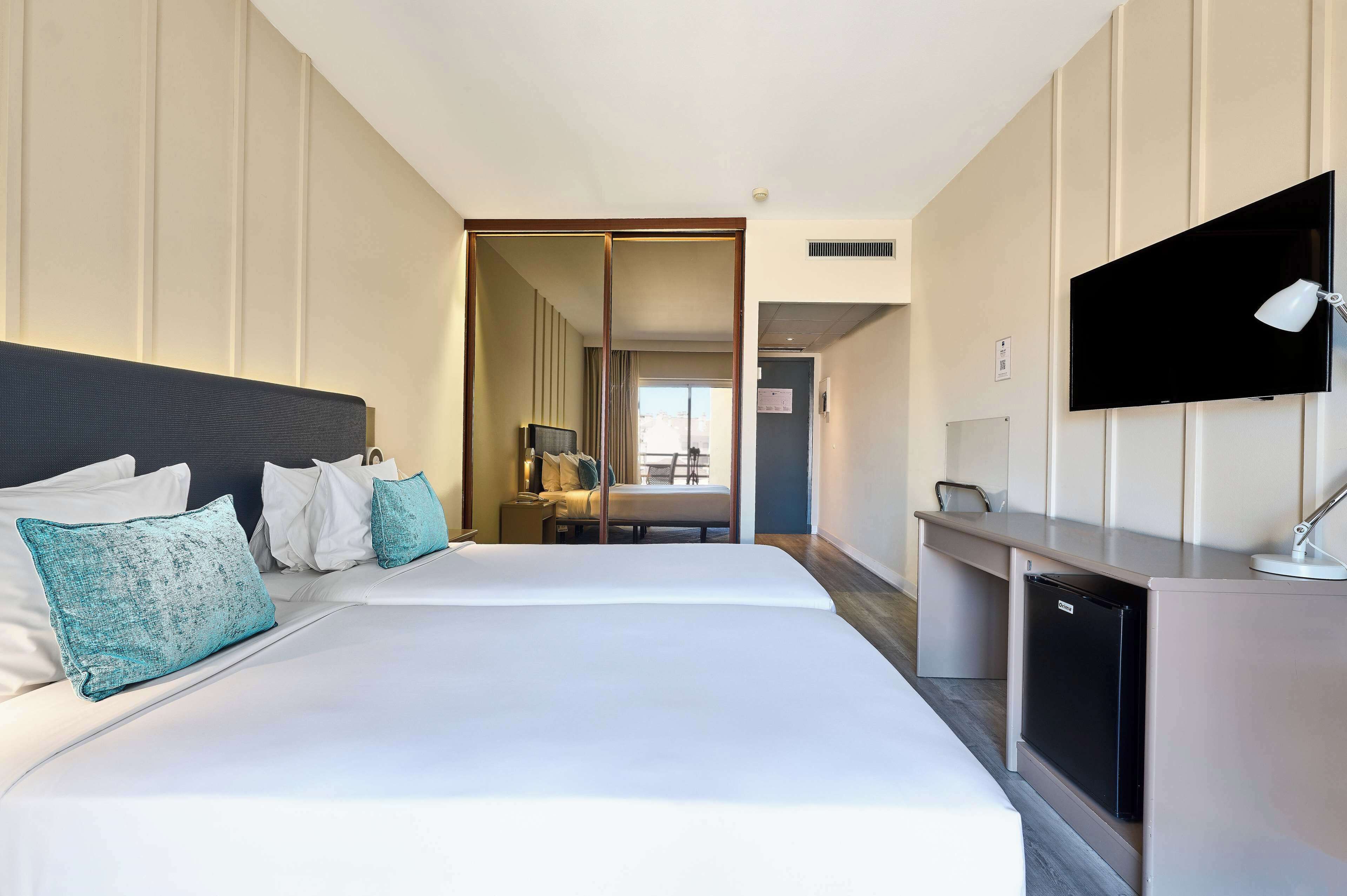 Quarto de hotel com duas camas, uma mesa, televisão e minibar.
