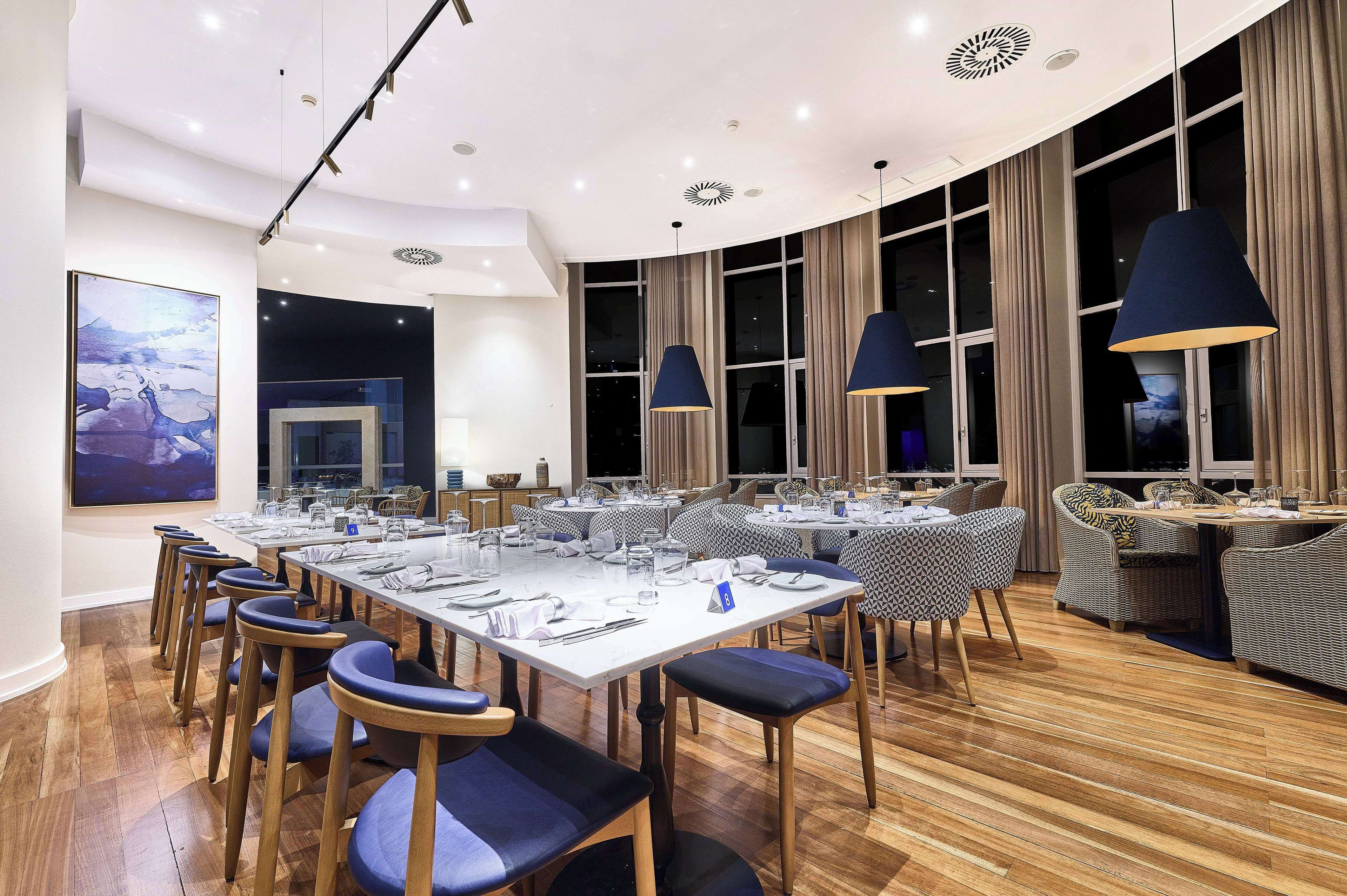 A modern dining area with a long table set for a meal, surrounded by chairs with blue cushions.