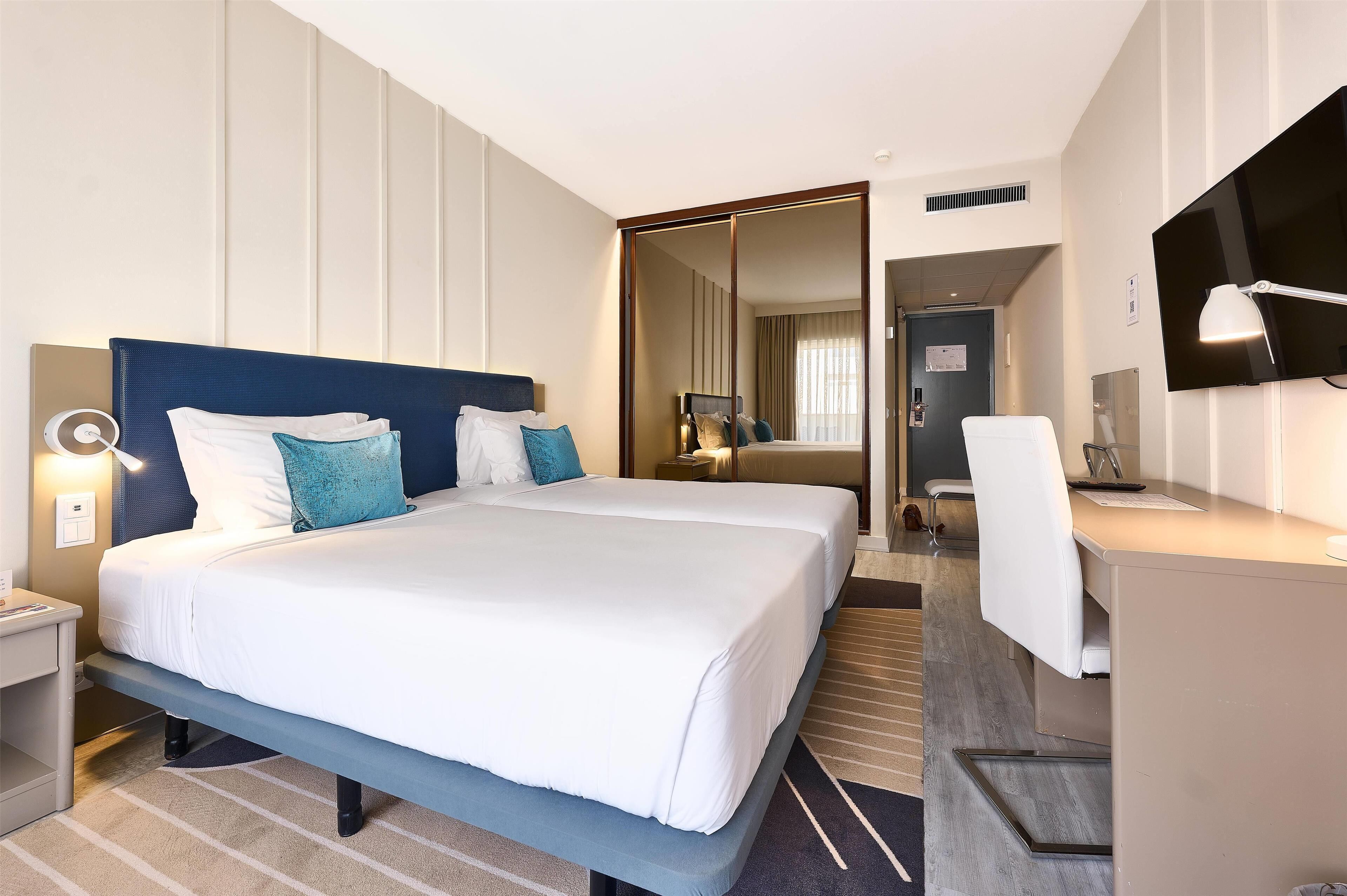 A hotel room with two beds, a desk, a chair, a TV, and a mirror.