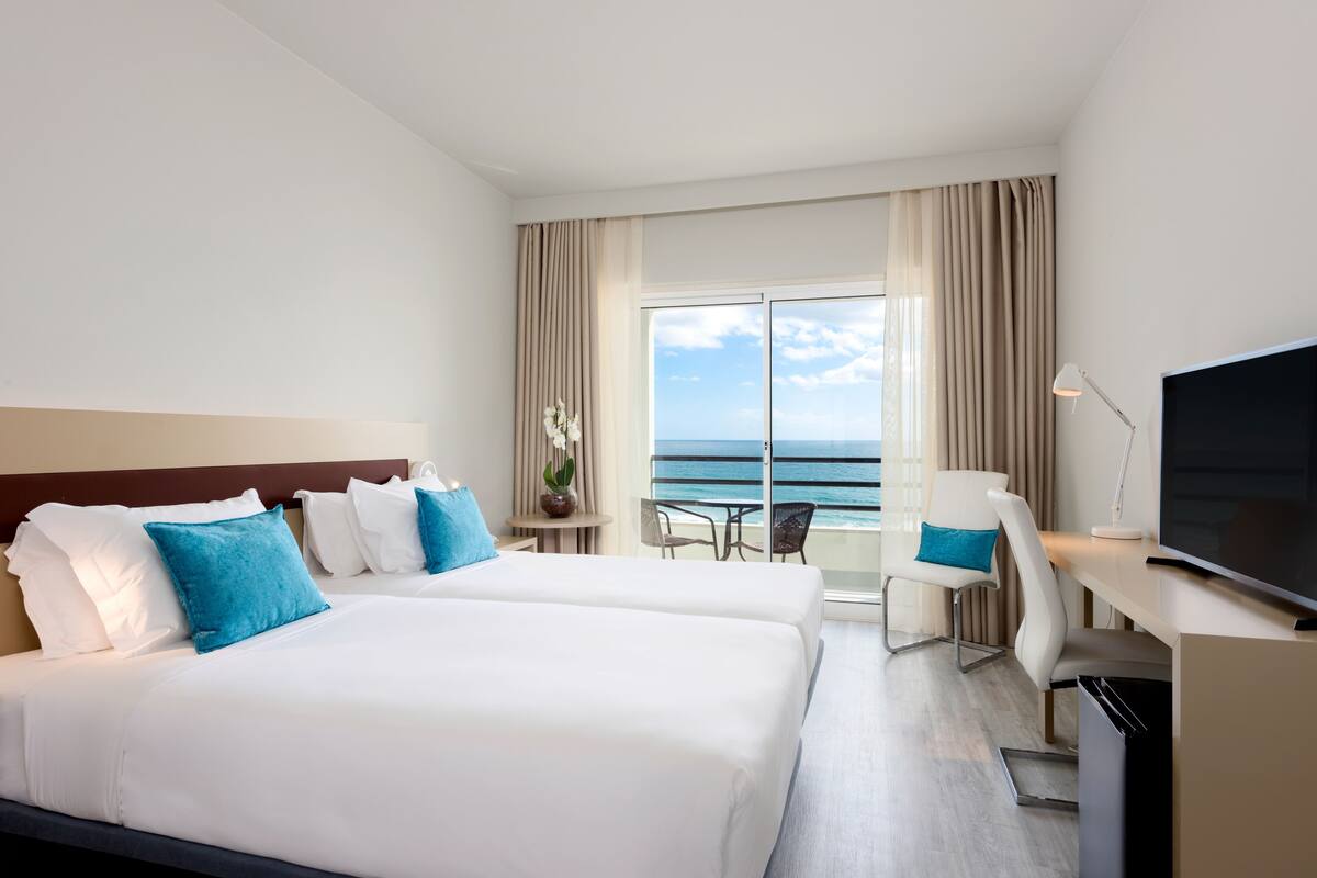 Habitación de hotel con dos camas, un escritorio, un televisor y un balcón con vista al océano.