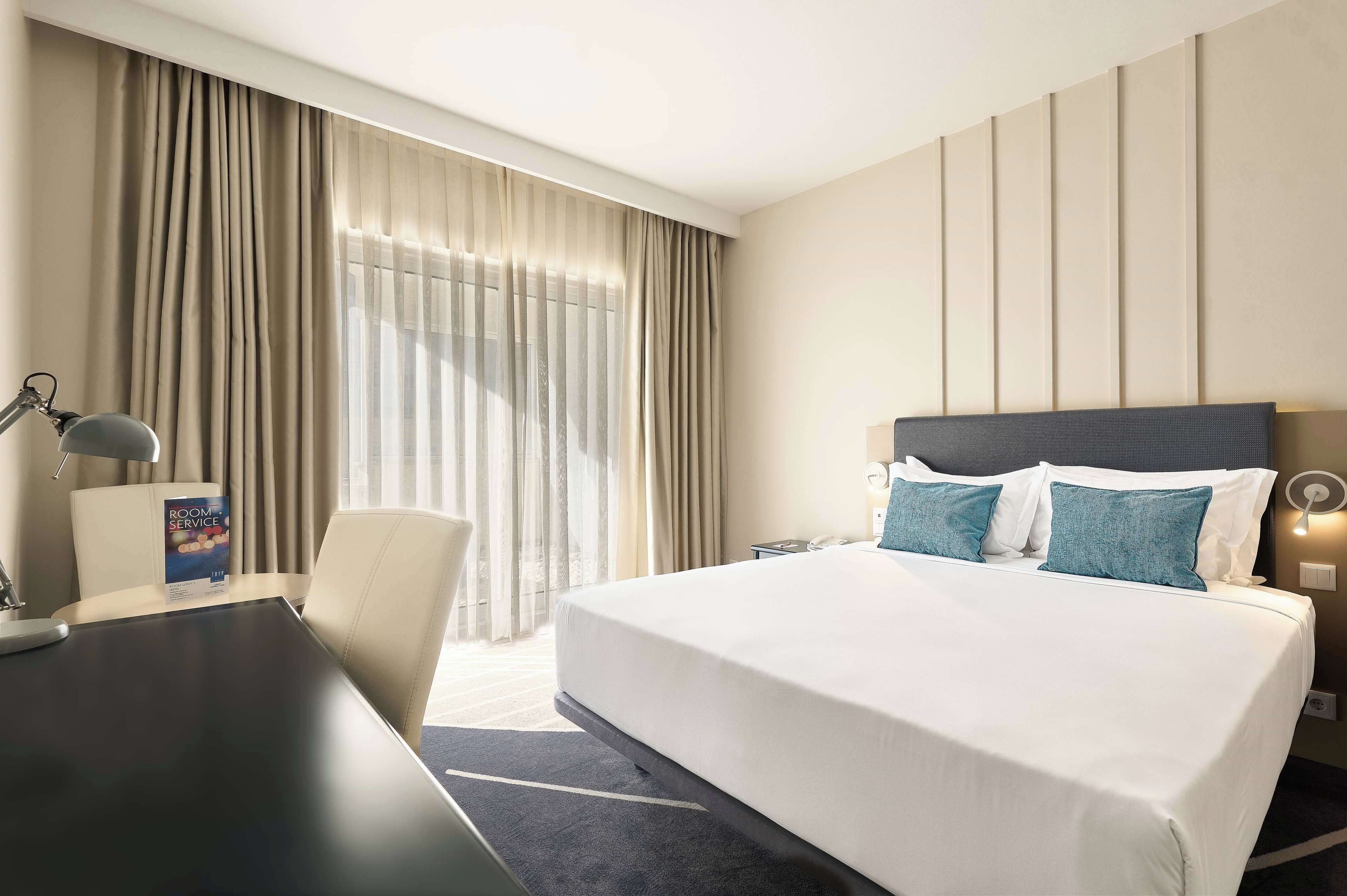 A hotel room with a large bed, a desk, and a chair. There is a window with curtains and a view of the outside.