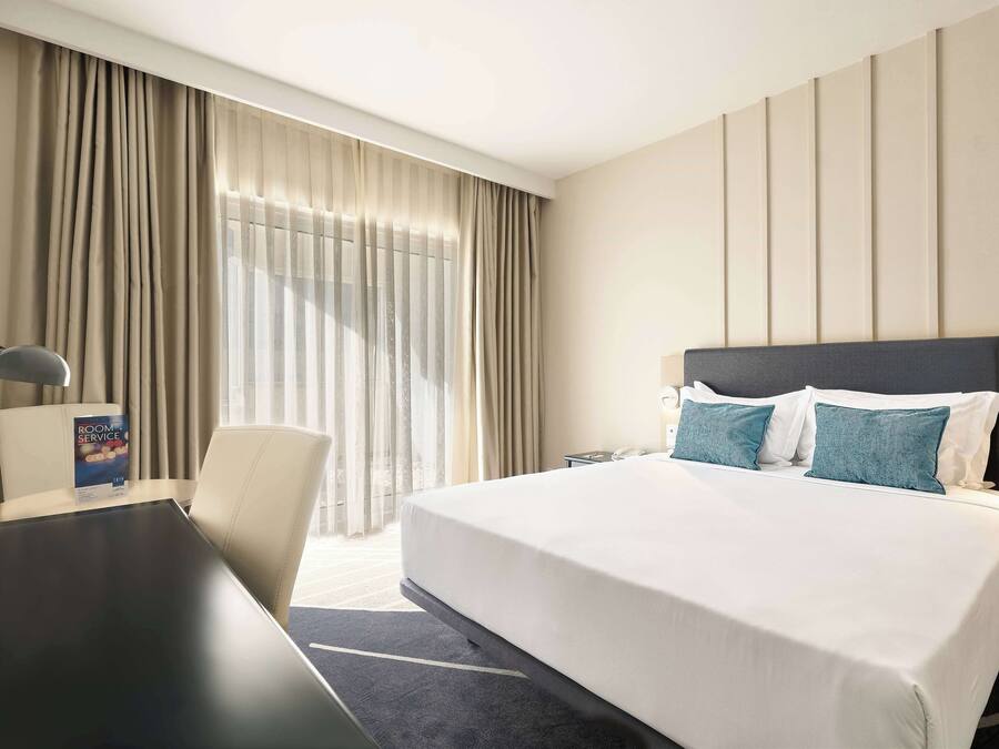 Ett hotellrum med en stor säng, ett skrivbord och en stol. Det finns ett fönster med gardiner och utsikt över omgivningen.