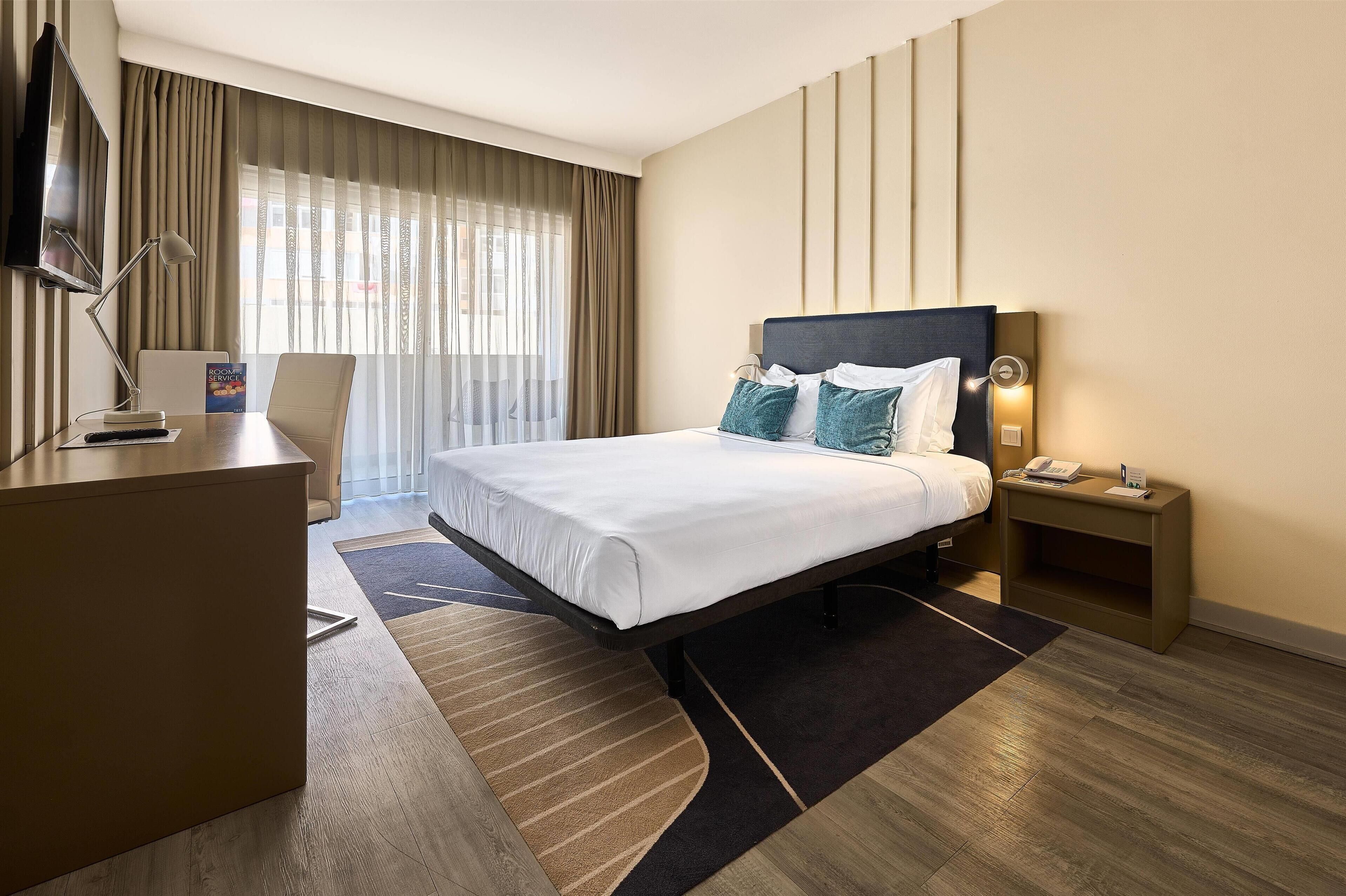 A hotel room with a bed, bedside table, desk, chair, and a TV.
