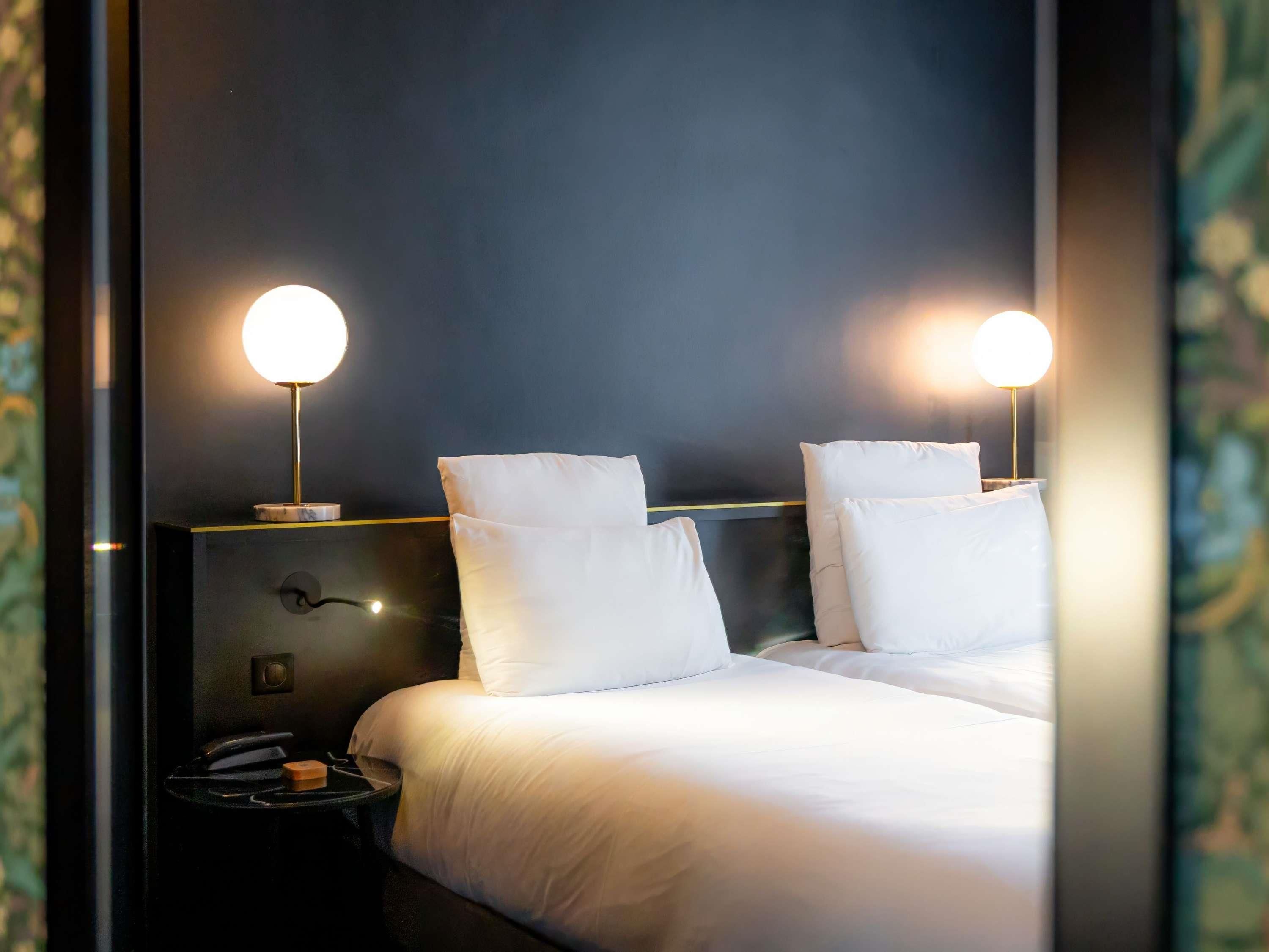 A hotel room with a bed, two bedside lamps, a nightstand, and a wall-mounted light switch.