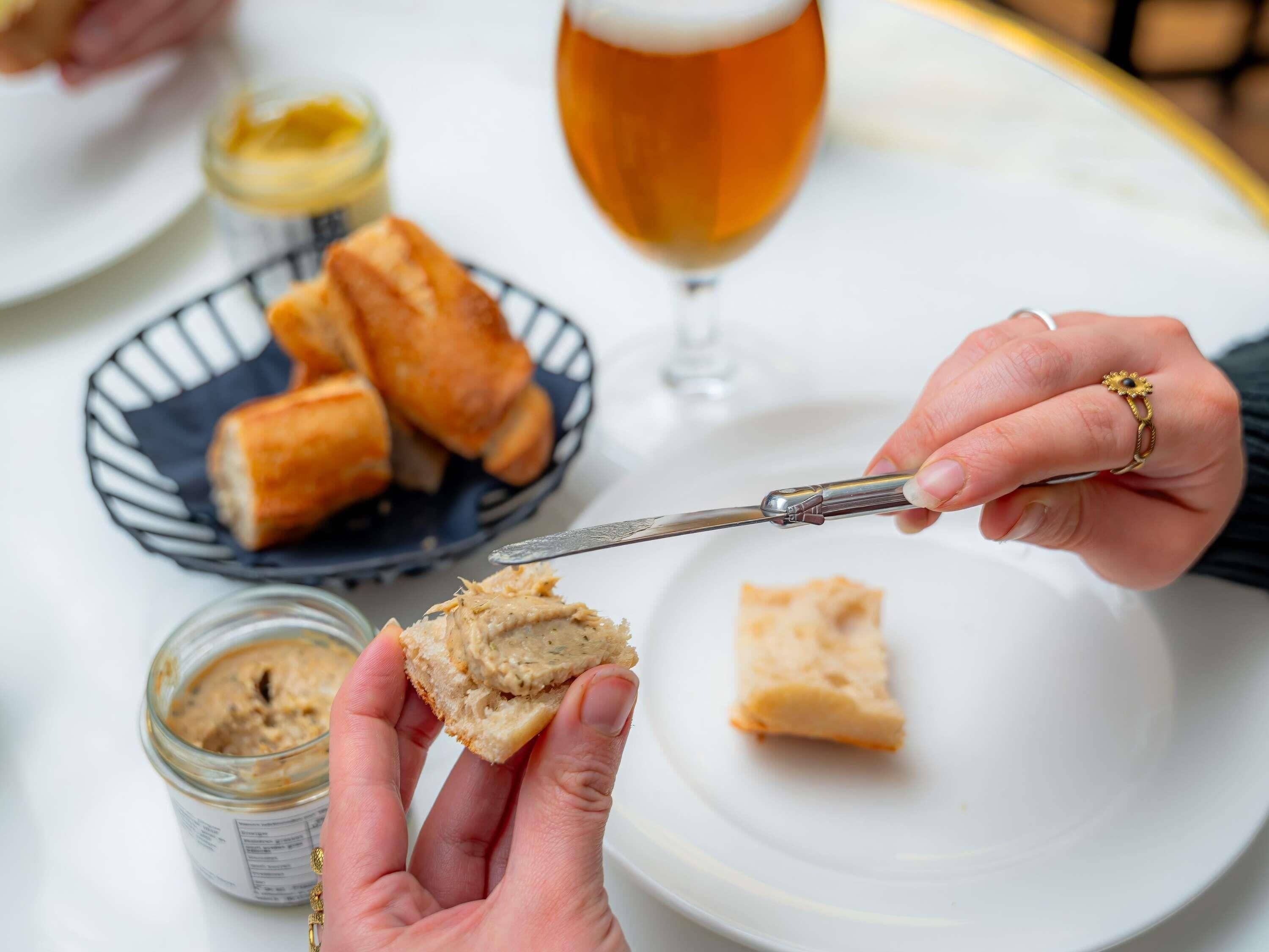 Una persona untando mantequilla en pan con un cuchillo sobre un plato blanco, con una jarra de cerveza y un bol de palitos de pan al fondo.