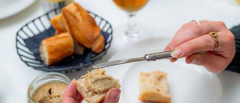 Una persona untando mantequilla en pan con un cuchillo sobre un plato blanco, con una jarra de cerveza y un bol de palitos de pan al fondo.